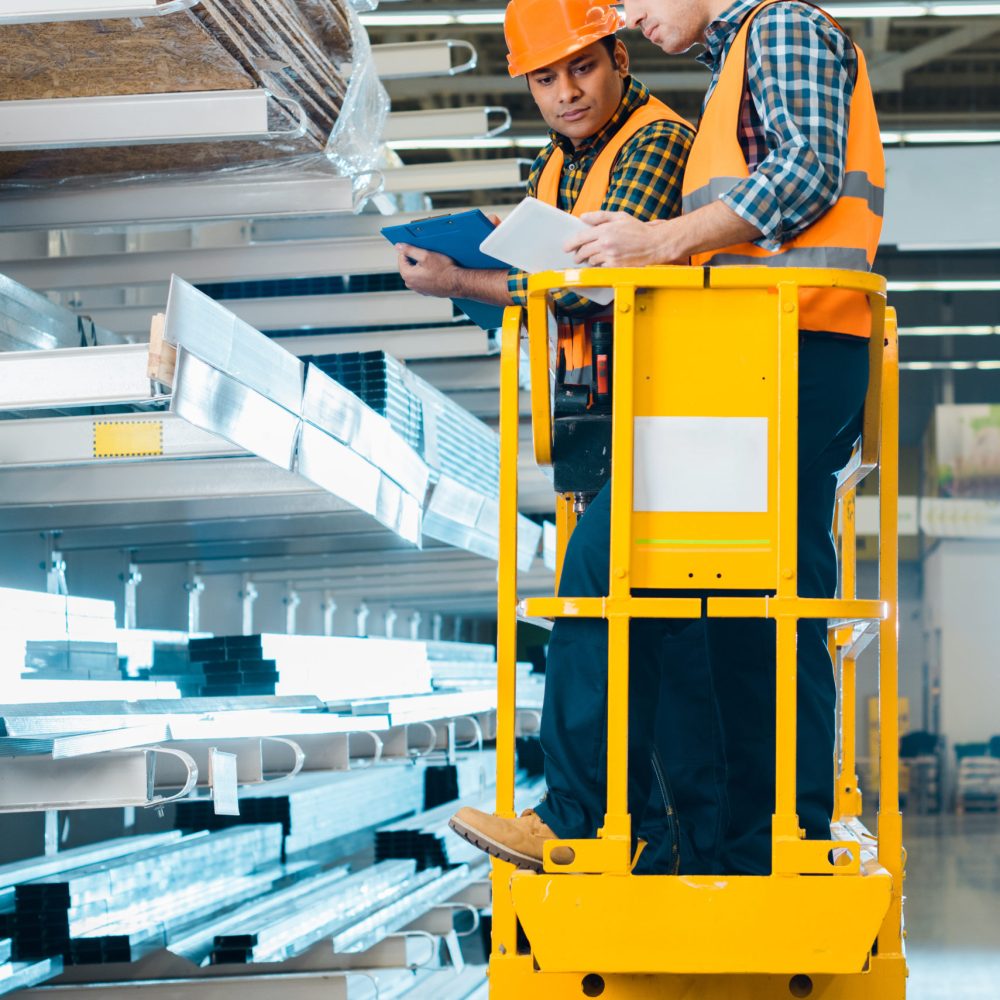 multicultural workers with digital tablet and clipboard standing on scissor lift near shelves with